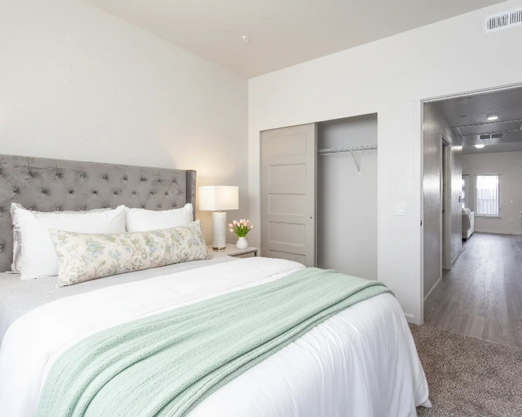 A neatly made bed with a green blanket and floral pillows in a softly lit bedroom, with a lamp and closed closet door nearby.