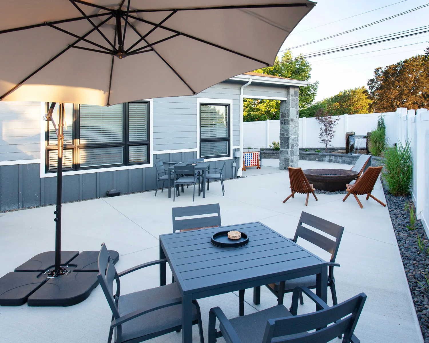 A modern outdoor patio with dark furniture, a large umbrella, and a fire pit surrounded by chairs in a fenced backyard.